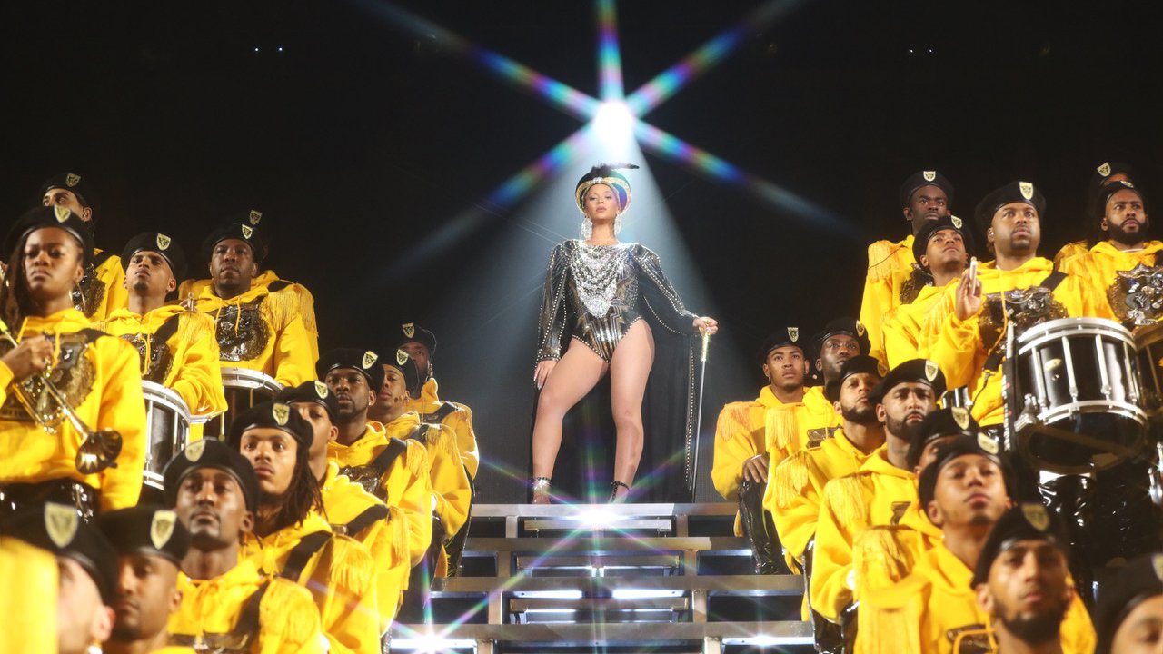 Beyonce stands onstage, surrounded by performers in yellow, in "Homecoming"