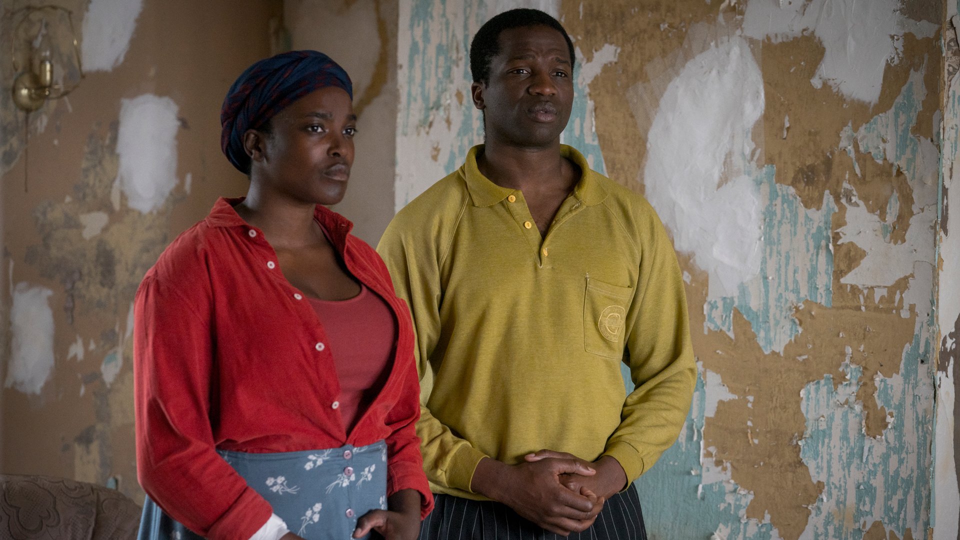 A woman in a red shirt and a man in a yellow shirt stand in a room with dilapidated walls.