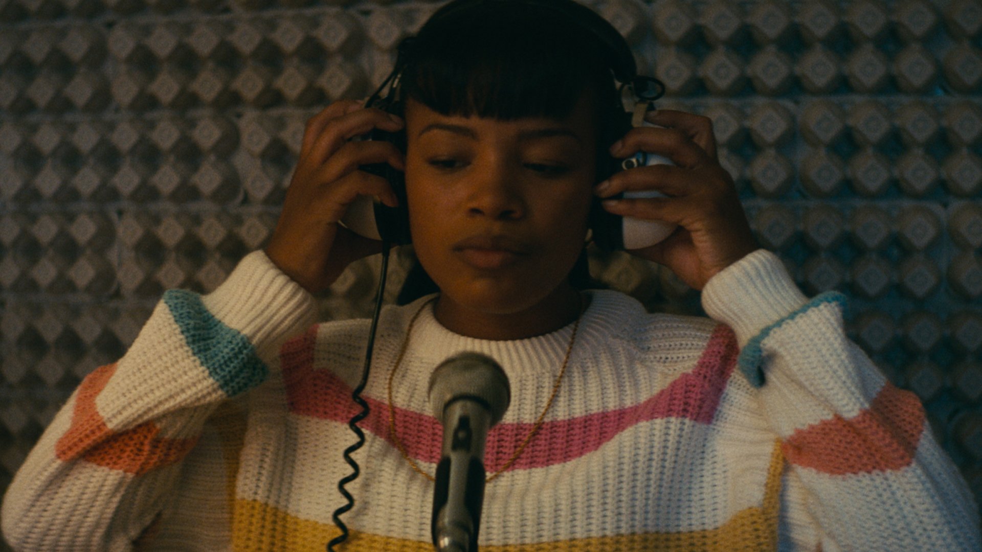 A woman in a striped sweater in front of a microphone puts headphones on and closes her eyes.