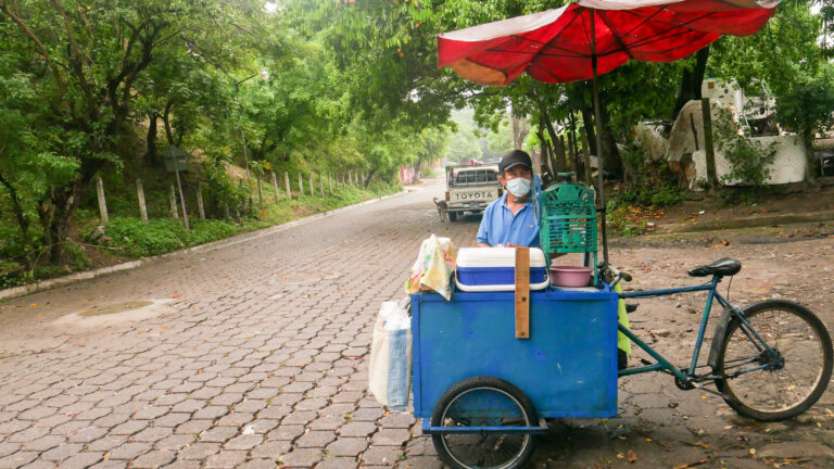 A vendor of a traditional ice cream in El Salvador, made with shaved ice bathed in fruit syrup, waits for customers on one of the streets of Parcelación El Ángel, in the municipality of Apopa, north of the capital. The locality is one of the epicenters where poor families have been organizing to block a residential development project, which will affect the local water supply and worsen the water shortage in the area. CREDIT: Edgardo Ayala/IPS