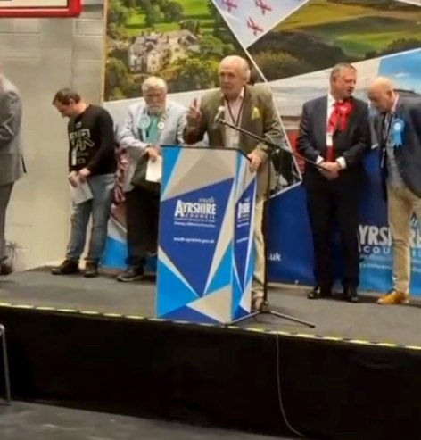 Wullie Hogg, an independent candidate, speaks at a podium on stage with other men during a council by-election.